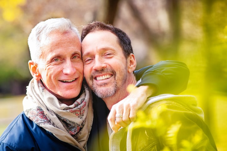 Couple of loving senior gay men portrait in a park in late October. They are both in hip mature fashion, happily and closely looking at the camera.