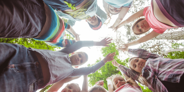 kids photographed from below gather in a circle, looking down at the camera, some with arms outstretched. a tree is in the background